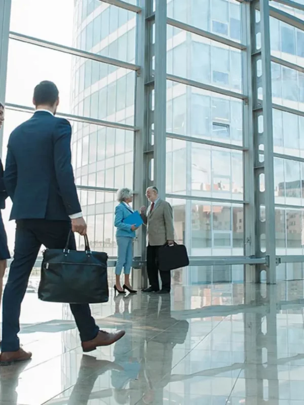 professionnels en tenue de travail se déplaçant dans un hall d’entreprise moderne avec de grandes fenêtres donnant sur une ville portuaire. Un homme et une femme au premier plan, en discussion tout en marchant, sont suivis de près par d’autres collègues. L’espace est lumineux et spacieux, reflétant une atmosphère de dynamisme et de collaboration dans un contexte professionnel.