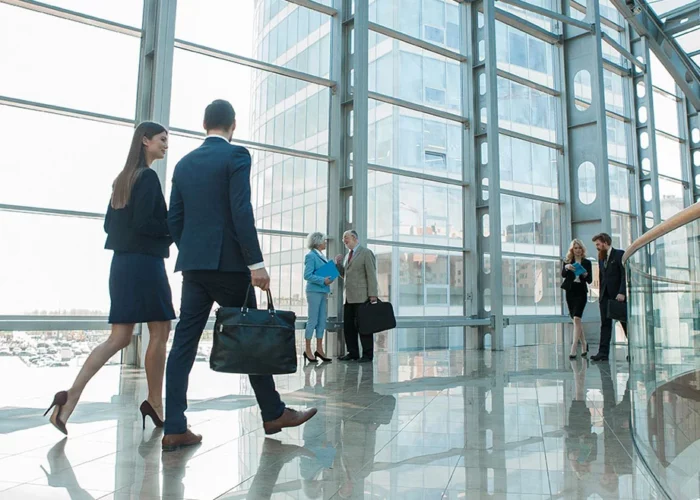 professionnels en tenue de travail se déplaçant dans un hall d’entreprise moderne avec de grandes fenêtres donnant sur une ville portuaire. Un homme et une femme au premier plan, en discussion tout en marchant, sont suivis de près par d’autres collègues. L’espace est lumineux et spacieux, reflétant une atmosphère de dynamisme et de collaboration dans un contexte professionnel.