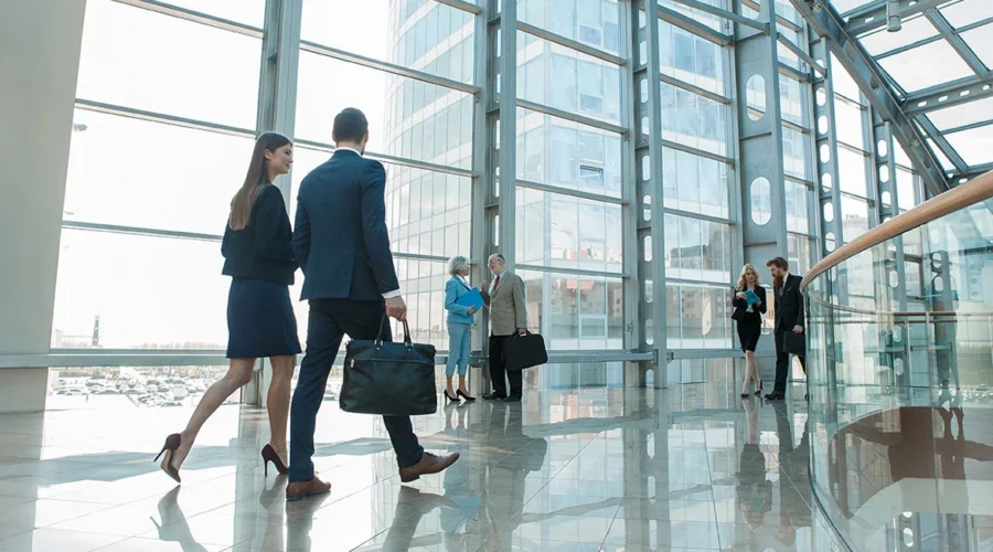 professionnels en tenue de travail se déplaçant dans un hall d’entreprise moderne avec de grandes fenêtres donnant sur une ville portuaire. Un homme et une femme au premier plan, en discussion tout en marchant, sont suivis de près par d’autres collègues. L’espace est lumineux et spacieux, reflétant une atmosphère de dynamisme et de collaboration dans un contexte professionnel.