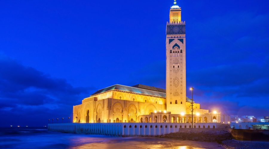 La mosquée Hassan II à Casablanca illuminée la nuit, avec son minaret dominant et orné qui se dresse contre un ciel bleu nuit. La structure imposante est située au bord de l’océan, avec des vagues floues visibles au premier plan. L’éclairage met en valeur les détails architecturaux sophistiqués et les motifs islamiques de la mosquée.