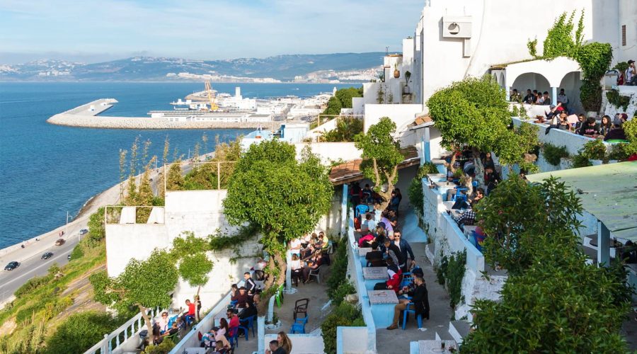 Vue sur un café en terrasse à Tanger, Maroc, avec des visiteurs assis à des tables bleues et blanches, profitant de la vue sur le port. Le café est niché sur une colline surplombant la mer, avec des bateaux et un large panorama urbain en arrière-plan sous un ciel clair.