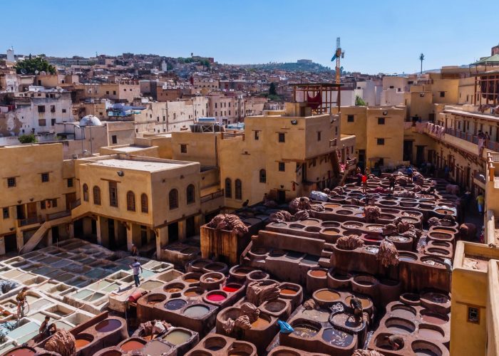 Vue aérienne des tanneries traditionnelles de Fès, Maroc, avec des ouvriers traitant le cuir dans des cuves colorées de teinture. Les bâtiments ocre et les maisons environnantes s’étendent jusqu’aux collines de la ville dense. Le ciel clair et la structure labyrinthique de la vieille ville ajoutent à l’atmosphère historique du site.