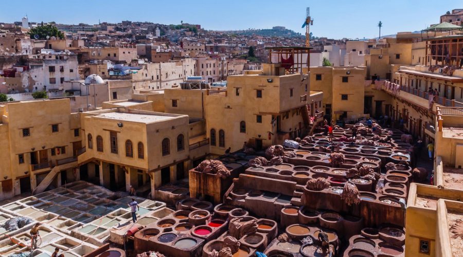 Vue aérienne des tanneries traditionnelles de Fès, Maroc, avec des ouvriers traitant le cuir dans des cuves colorées de teinture. Les bâtiments ocre et les maisons environnantes s’étendent jusqu’aux collines de la ville dense. Le ciel clair et la structure labyrinthique de la vieille ville ajoutent à l’atmosphère historique du site.