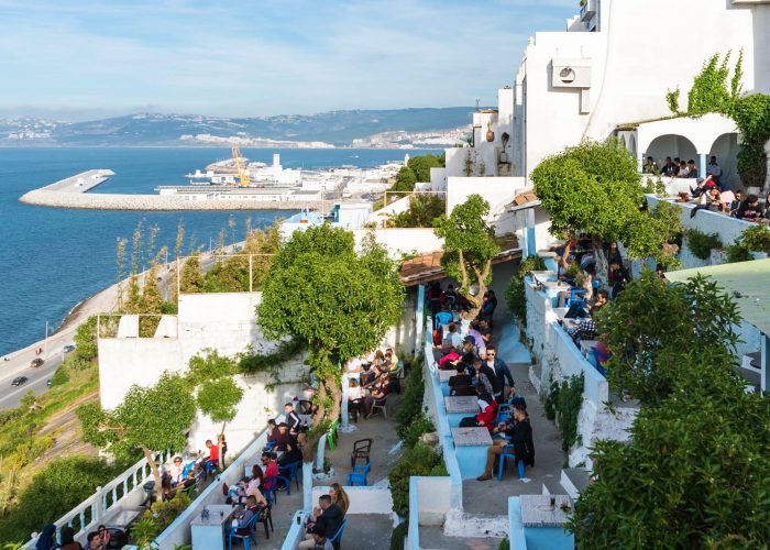 Vue sur un café en terrasse à Tanger, Maroc, avec des visiteurs assis à des tables bleues et blanches, profitant de la vue sur le port. Le café est niché sur une colline surplombant la mer, avec des bateaux et un large panorama urbain en arrière-plan sous un ciel clair.