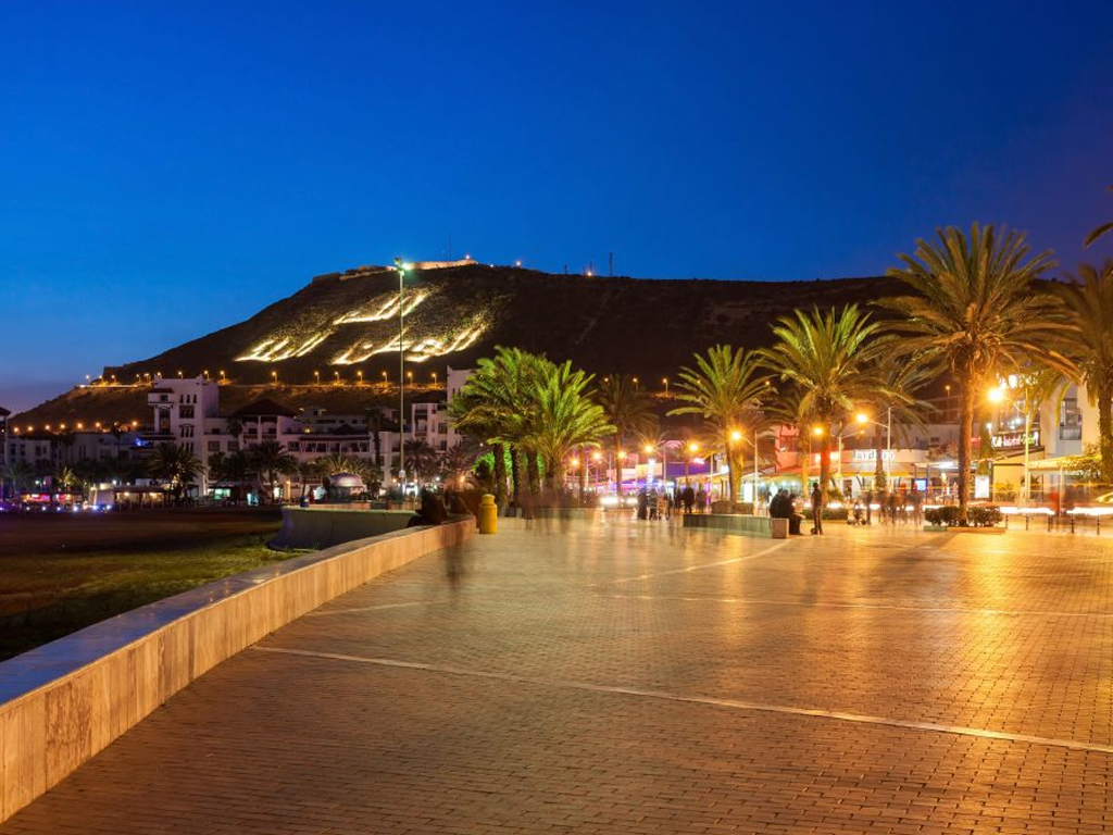 Promenade en bord de mer à Agadir illuminée la nuit avec la montagne portant l’inscription ‘Allah, Al-Watan, Al-Malik’ en arrière-plan.