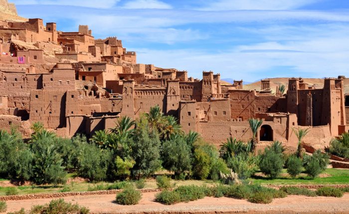 Vue panoramique de la kasbah d’Ait Ben Haddou au Maroc, un village fortifié traditionnel composé de structures en pisé. Les bâtiments ocre avec des tours fortifiées et des murs crénelés se dressent sur fond de montagnes arides. Le premier plan montre un lit de rivière asséché bordé de végétation luxuriante, notamment des palmiers et des buissons.