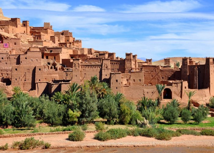 Vue panoramique de la kasbah d’Ait Ben Haddou au Maroc, un village fortifié traditionnel composé de structures en pisé. Les bâtiments ocre avec des tours fortifiées et des murs crénelés se dressent sur fond de montagnes arides. Le premier plan montre un lit de rivière asséché bordé de végétation luxuriante, notamment des palmiers et des buissons.