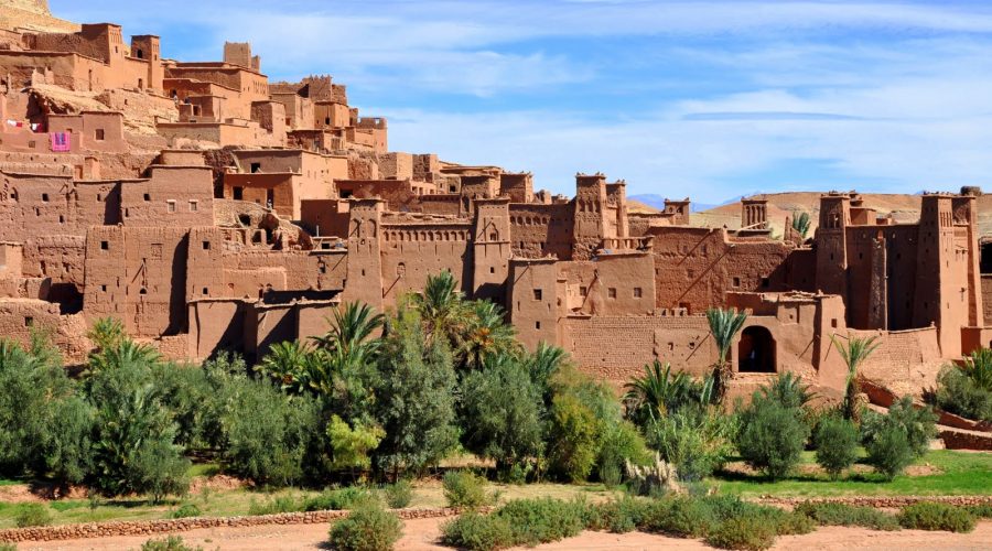 Vue panoramique de la kasbah d’Ait Ben Haddou au Maroc, un village fortifié traditionnel composé de structures en pisé. Les bâtiments ocre avec des tours fortifiées et des murs crénelés se dressent sur fond de montagnes arides. Le premier plan montre un lit de rivière asséché bordé de végétation luxuriante, notamment des palmiers et des buissons.