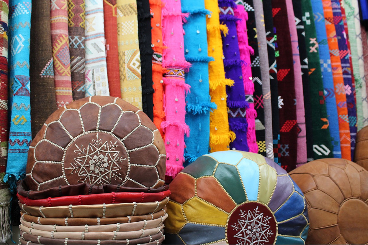 Étals de marché marocain présentant une variété de tapis colorés tissés à la main accrochés en arrière-plan, avec des poufs en cuir traditionnels ornés de motifs brodés en premier plan.