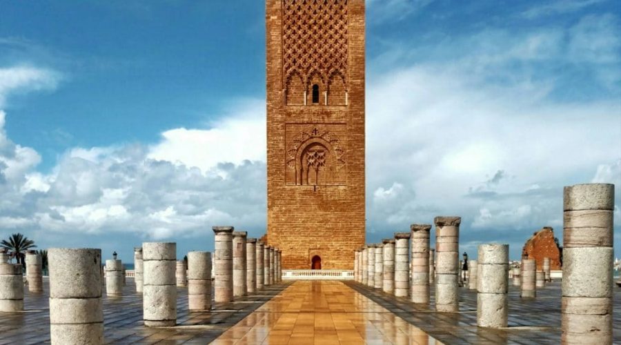 Vue de la Tour Hassan à Rabat, Maroc, sous un ciel nuageux. Au premier plan, un grand lustre ornemental en métal doré avec des détails ajourés repose sur une base en marbre. L’arrière-plan montre la célèbre tour en brique rouge, incomplète, entourée de colonnes blanches éparpillées sur une vaste esplanade.