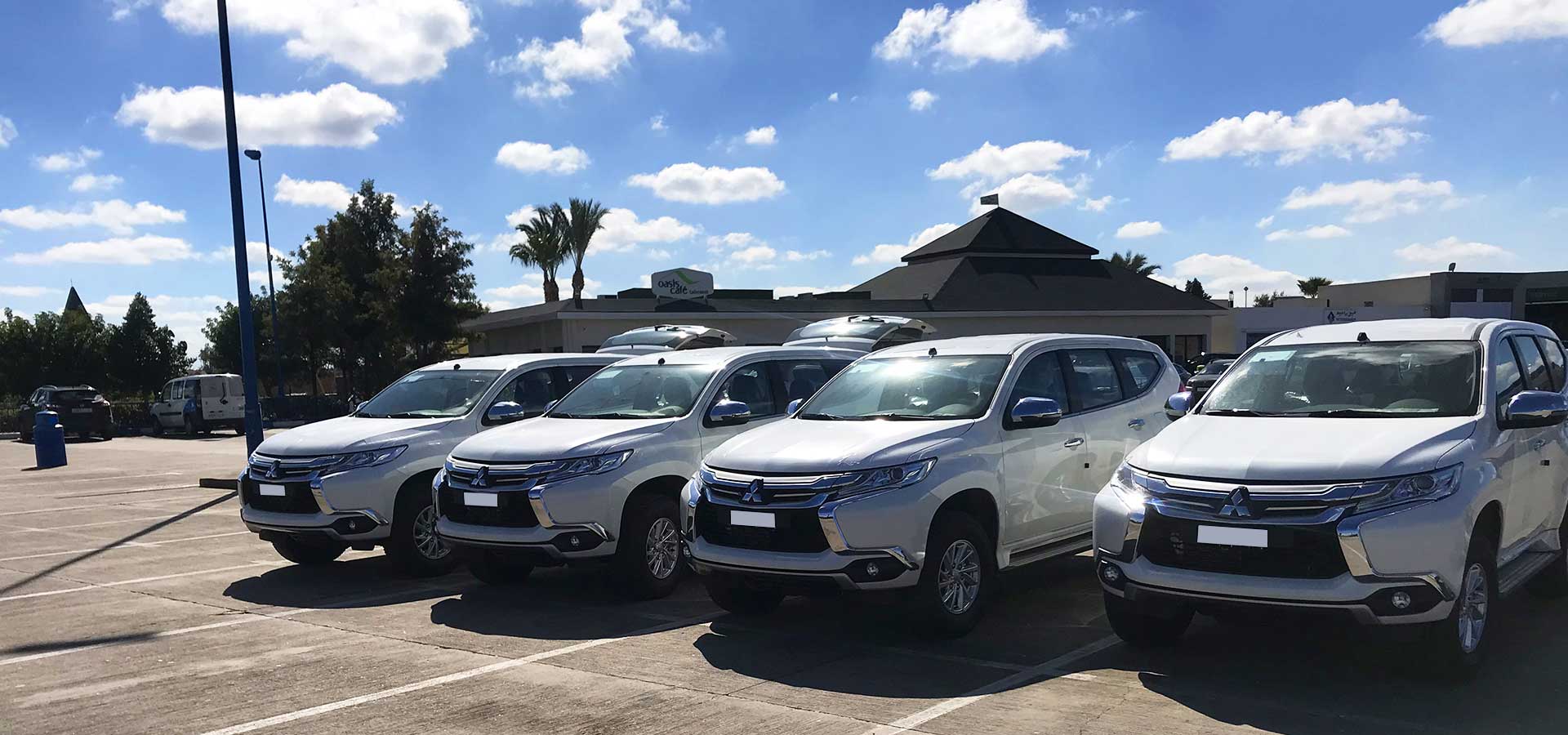 Alignement de plusieurs SUV Mitsubishi blancs garés sur un parking en plein air, sous un ciel bleu avec quelques nuages.