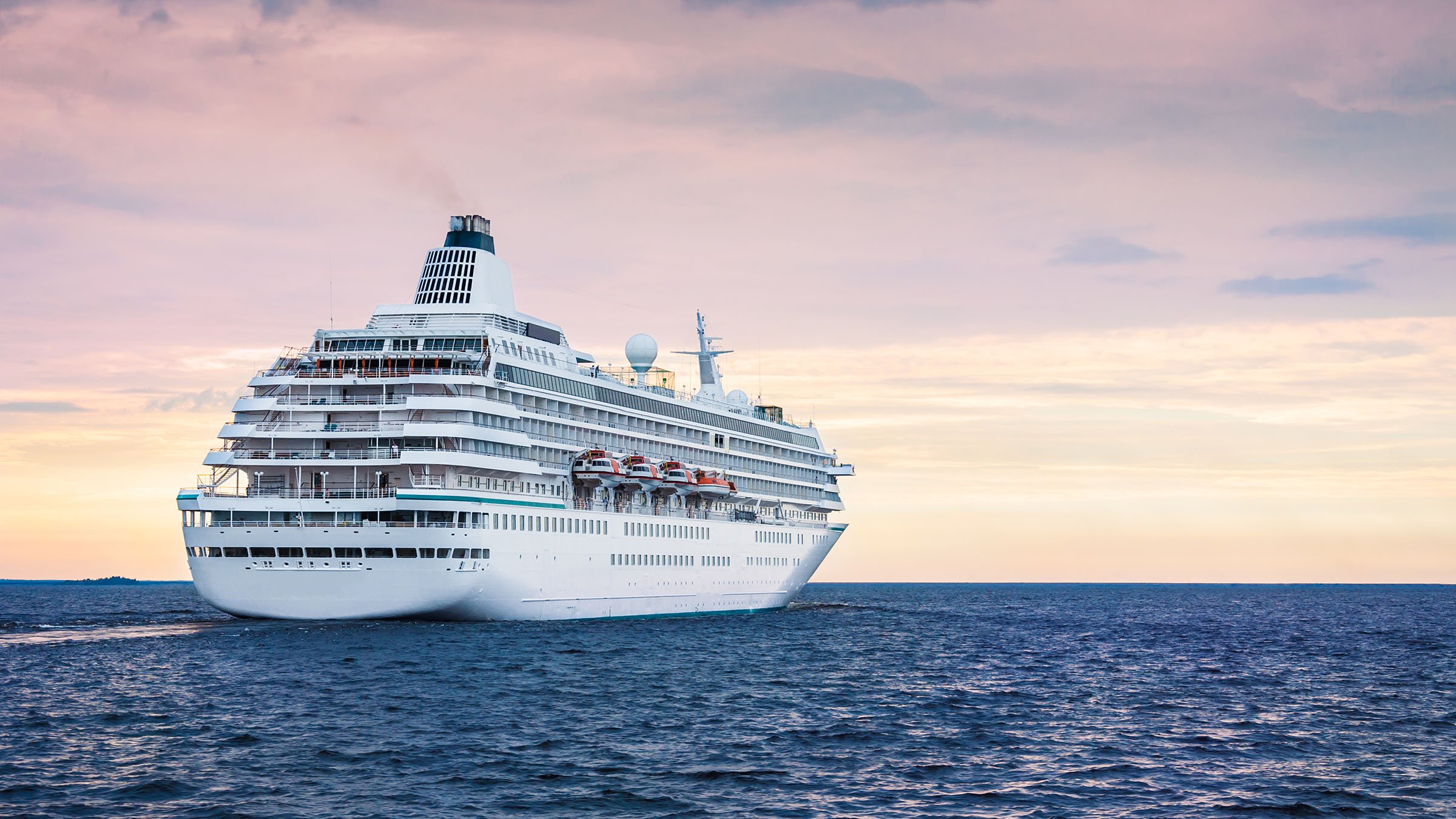 Grand navire de croisière blanc naviguant sur l’océan sous un ciel pastel au coucher du soleil.