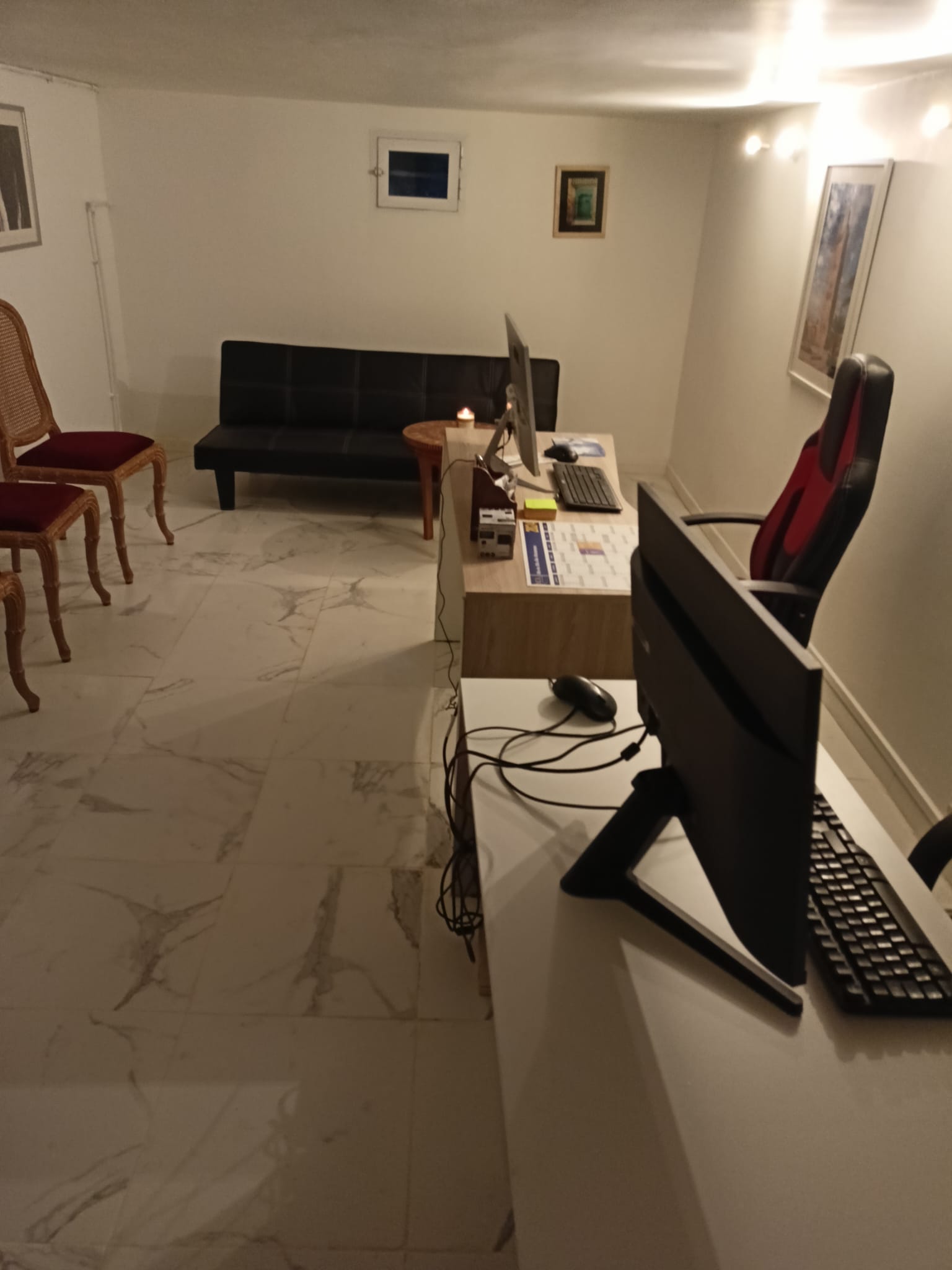 Intérieur d’un bureau avec un sol en marbre blanc veiné. À gauche, une table en bois avec deux chaises, une classique en velours rouge et une autre en bois traditionnel. À droite, un espace de travail avec un bureau blanc équipé d’un ordinateur, un écran, un clavier, une souris, et un fauteuil de bureau ergonomique rouge et noir. Un canapé noir se trouve en arrière-plan, sous deux tableaux accrochés sur le mur blanc.