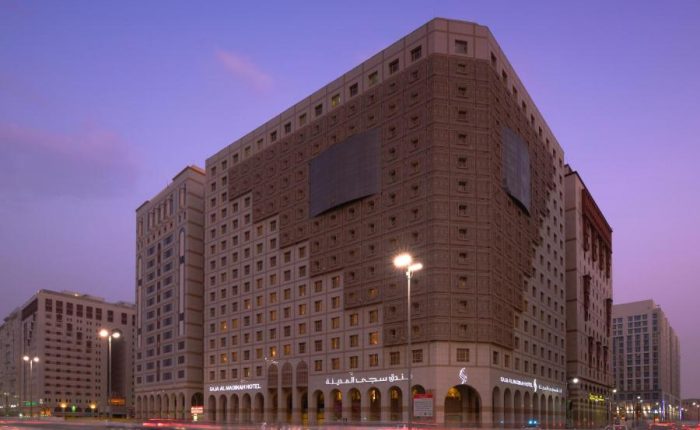 Vue crépusculaire d’un grand hôtel urbain moderne, le Salam Madinah Hotel, au crépuscule. L’architecture de l’hôtel est caractérisée par un large bâtiment carré avec des motifs de fenêtres répétitifs et un grand panneau publicitaire vide. En premier plan, les lumières des véhicules passant à grande vitesse sur une route urbaine créent des traînées lumineuses, ajoutant un effet dynamique à la scène.