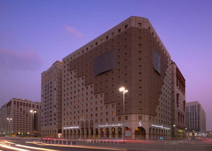 Vue crépusculaire d’un grand hôtel urbain moderne, le Salam Madinah Hotel, au crépuscule. L’architecture de l’hôtel est caractérisée par un large bâtiment carré avec des motifs de fenêtres répétitifs et un grand panneau publicitaire vide. En premier plan, les lumières des véhicules passant à grande vitesse sur une route urbaine créent des traînées lumineuses, ajoutant un effet dynamique à la scène.