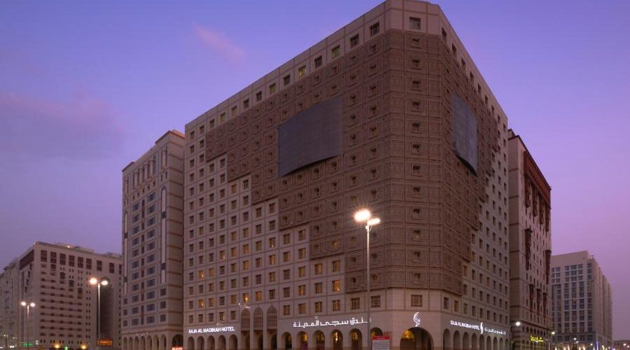 Vue crépusculaire d’un grand hôtel urbain moderne, le Salam Madinah Hotel, au crépuscule. L’architecture de l’hôtel est caractérisée par un large bâtiment carré avec des motifs de fenêtres répétitifs et un grand panneau publicitaire vide. En premier plan, les lumières des véhicules passant à grande vitesse sur une route urbaine créent des traînées lumineuses, ajoutant un effet dynamique à la scène.