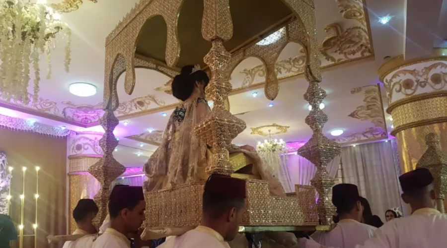 mariée marocaine assise dans une amariya, une structure dorée richement ornée portée par plusieurs hommes vêtus de tenues traditionnelles. La scène est illuminée par des lumières de cérémonie et un décor somptueux aux accents dorés et mauves, typique des mariages marocains.